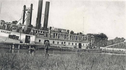 Black and white photo of large steamship on the river, people watching on the bank