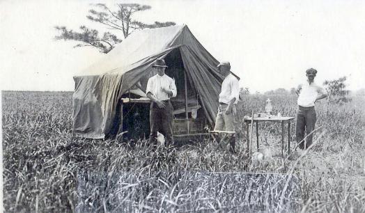 Black and white photo of men in a field with a tent and table set up